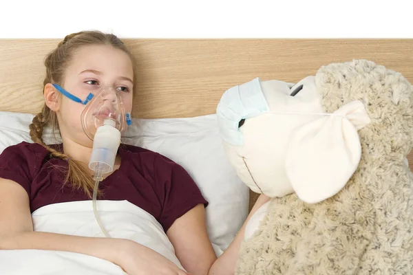 girl with inhaler mask - respiratory problems for asthma. girl with a mask inhaler lies with a toy in bed and breathes adrenaline. health care and sick child concept