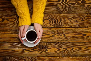 Fotokopi alanı olan eski ahşap masa manzaralı kahve fincanı. Sıcak, sarı ev kazağı giymiş bir kadın elinde bir kupa kahveyle içkinin tadını çıkarıyor..