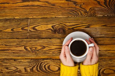 Fotokopi alanı olan eski ahşap masa manzaralı kahve fincanı. Sıcak, sarı ev kazağı giymiş bir kadın elinde bir kupa kahveyle içkinin tadını çıkarıyor.
