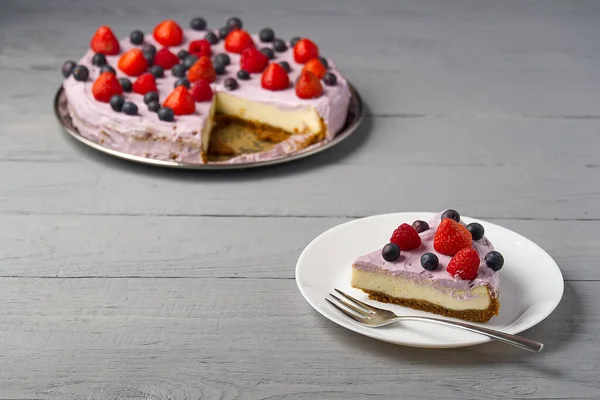 Rebanada de pastel de queso hecho en casa con bayas en el plato. Tarta de queso con fresa, arándano y frambuesa sobre fondo de madera gris . — Foto de Stock