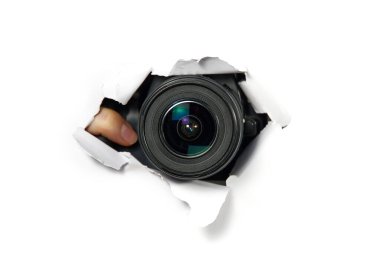 Beyaz kağıttaki delikten dışarı bakan telefoto lensli siyah kamera. Paparazzi kavramı, casusluk, sarı basın. Kamera merceği duvardaki delikten bakıyor.