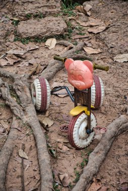 An old, ruined lone and left and child tricycle with a pink bear head among the roots of a spung tree in Cambodia clipart