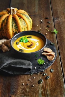 Zencefilli baharatlı balkabağı çorbası, siyah kavanozda sunulur kayrak taşı üzerinde balkabağı yağı, krema ve balkabağı çekirdekli ahşap masa. 