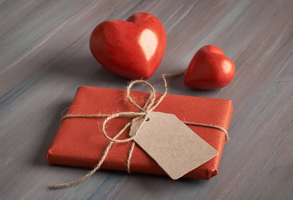 Packed present,  wrapped in red paper with brown cord and blank paper tag, and two red stone hearts on rustic wooden background. Valentines or Mother\'s day concept. Space for your text on the tag.