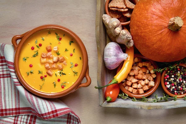 Pumpkin soup in ceramic bowl on light wood served with thyme and croutons. Next to it is a tray with healthy soup ingredients. Flat lay, view from above.