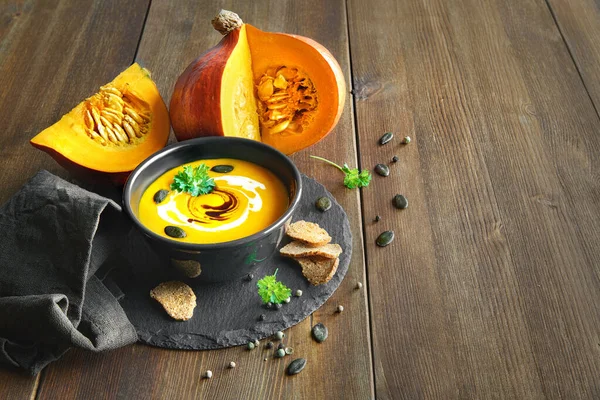 Spicy pumpkin soup with ginger, served in black jar on a slate stone with pumpkin oil, cream and pumpkin seeds on the wooden table. Space for your text.