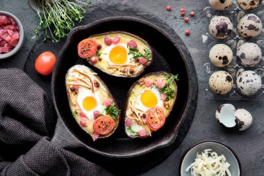 Keto diyet yemeği: jambon küplü avokado tekneleri, bıldırcın yumurtaları, peynir ve kiraz domatesleri demir tavada havluyla, üstü koyu arkaplanda malzemelerle kaplı.