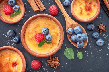 Creme brulee (krem brulee, yanmış krema) ahududu, yabanmersini ve nane şekeri ile terakota kil pişirme kabında. Üst görünüm, koyu taş arkaplan.