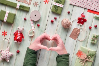 Çeşitli Noel ya da yeni yıl tatillerinde çevre dostu dekorasyonlar, kağıt paketler ve el yapımı veya sıfır atık hediye. Tahtaya yaslanmış, elleri yeşil kollu, kalp işareti var..