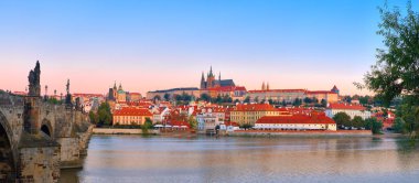 Romantik Prag 'ın şafak vakti panoramik görüntüsü. Güneşin doğuşunda Vltava nehrinin kenarında, solunda Charles Köprüsü ve St. Vitus katedrali ve kalesi ile tarihi gökyüzü.. 