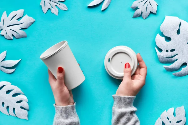 Stylish reusable eco coffee travel mug, bamboo cup with lid in hands. Blue mint aqua menthe paper background, cut out exotic leaves. Zero waste eco friendly lifestyle trend.