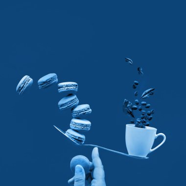 Monochrome blue color toned image. Balancing cup of coffee and macaroons on index finger. Perfect balance concept. Creative square food composition on dark blue square paper background, copy space. clipart