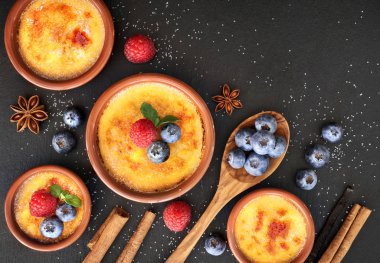 Creme brulee (krem brulee, yanmış krema) ahududu, yabanmersini ve nane şekeri ile terakota kil pişirme kabında. Üst görünüm, koyu taş arkaplan.