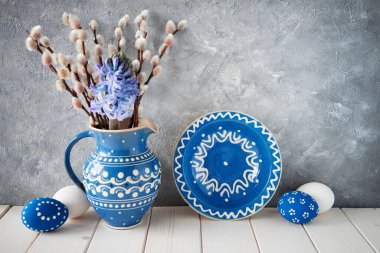 Mavi seramik sürahide bir sürü kedi söğüdü, eşleşen tabaklar, Paskalya yumurtaları ve gri arka planda bahar süslemeleri.