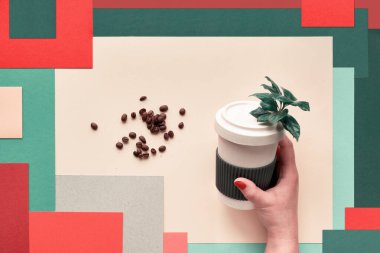 Bambu kahve fincanı (fincanı veya seyahat kupası) soyut geometrik katmanlı çok renkli dikdörtgen kağıdın arka planında kadın ellerinde bulunur. Yaratıcı düzlük, üst görünüm, çevre dostu sıfır atık bardak.