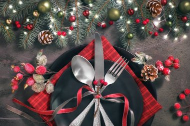 Karanlık arka planda bir Noel masası. Yeşil ve kırmızı renkli Xmas dekorasyonları, buzlu böğürtlen, incik boncuk, siyah tabak ve çanak çömlek..