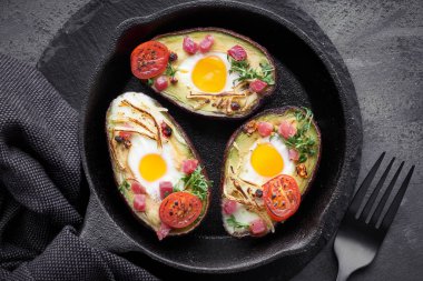 Keto diyet yemeği: Jambon küplü avokado tekneleri, bıldırcın yumurtaları, peynir ve tere lahanaları dökme demir tavada, koyu arkaplan havlusu, üst görünüm