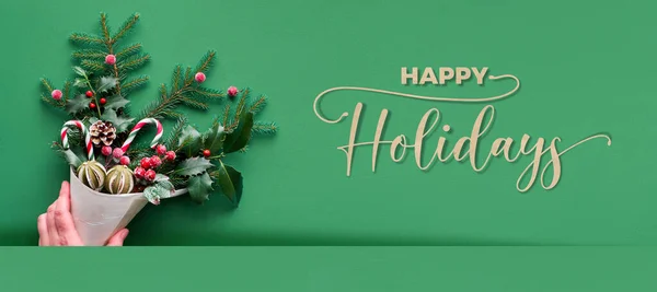 クリスマスのパノラマの背景フラットテキスト ハッピーホリデー と緑の紙の上に横たわっていた キャンディーの缶やベリーで装飾されたモミやホリー小枝とベニヤコーンを保持手 パノラマ構成 — ストック写真