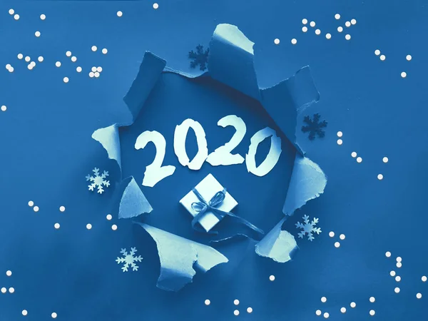 Trnedy classic blue color paper flat lay with snowflakes and ripped hole in the middle. There are New Year\'s gift and paper number 2020 in the hole. Happy New Year 2020!