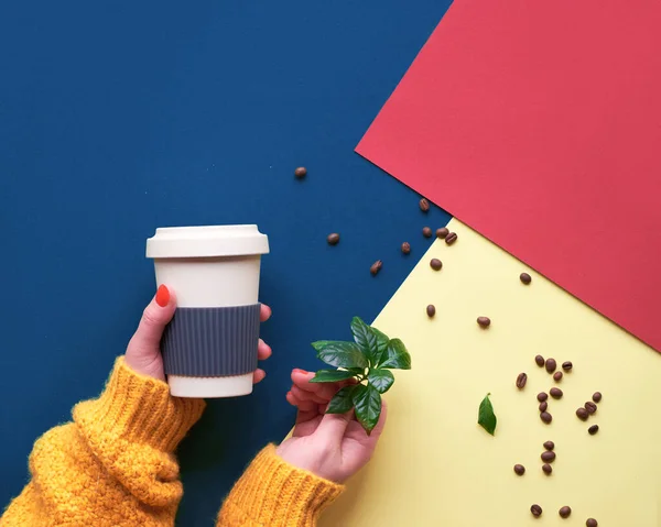 Zero waste coffee concept. Geometric top view on split three tone paper, red, blue and yellow.Eco friendly reusable coffee cups, hands in orange sweater holding the mug and coffee plant.