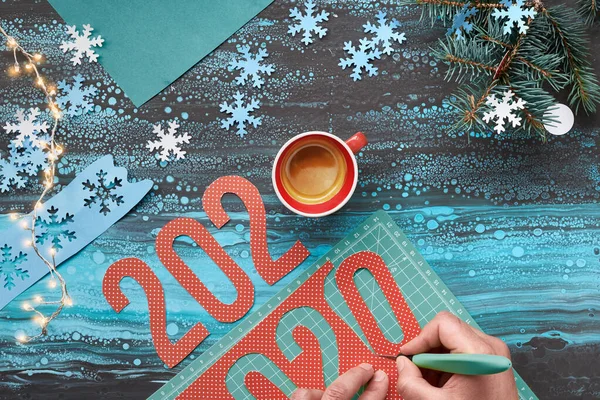 Concept winter background, paper craft with hand cutting number 2020, cup of coffee and Christmas decorations. Flat lay, top view on abstract mint color background.