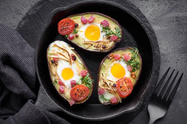 Keto diyet yemeği: Jambon küplü avokado tekneleri, bıldırcın yumurtaları, peynir ve tere lahanaları dökme demir tavada, koyu arkaplan havlusu, üst görünüm