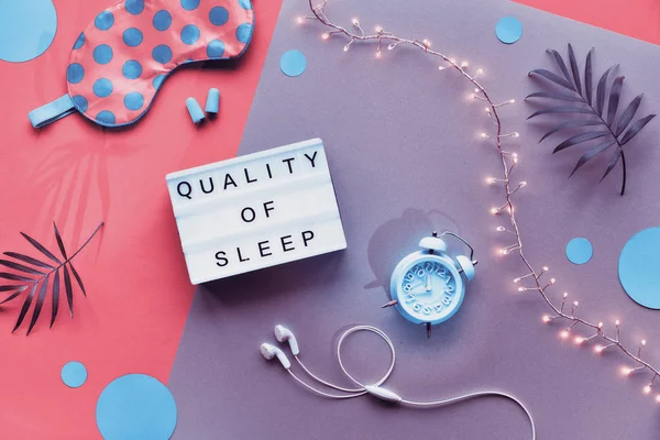 Lightbox with text "Quality of sleep". Healthy night sleep creative flat lay. Sleeping mask, blue mint alarm clock, earphones, earplugs. Split pink and silver background with circles and palm leaves — ストック写真