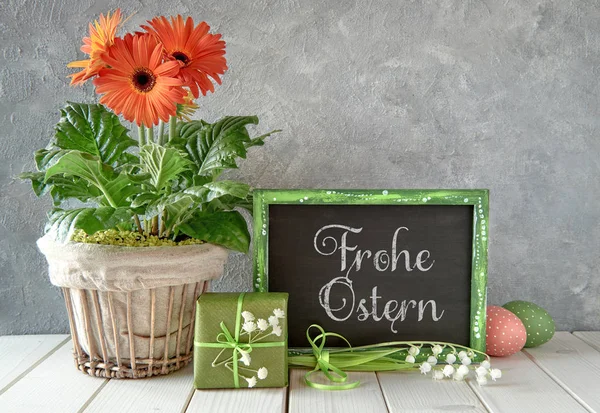 Happy Easter! Text on chalk board, orange gerbera flowers in flower pot and green gift box with baby breath flowers and lily of the valley on rustic table, Easter decorations
