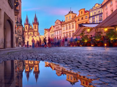 Prag 'daki Aziz Mary Tyn Kilisesi' nde yaz yağmurundan sonra su birikintisine yansıyan yansımayla Eski Şehir Meydanı 'na doğru yürüyen turistler görülüyor. Romantik Prag, gün batımında seyahat geçmişi.