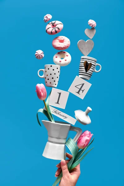 Día de San Valentín fondo divertido en menta azul. Fecha 14 de febrero en el calendario de madera con tazas de café, tulipán, rosquillas y malvaviscos. Mano femenina sosteniendo pirámide de equilibrio en la cafetera . — Foto de Stock