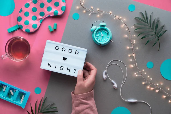 テキスト"おやすみ"を手にライトボックス。健康な夜の睡眠創造的なフラットレイアウト。睡眠マスク、ブルーミント目覚まし時計、イヤフォン、耳栓。円とヤシの葉を持つ2つのトーンピンクの銀の背景. — ストック写真
