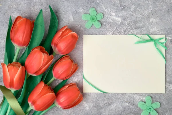 Spring greetings, flat lay with red tulips and blank paper card on grey textured background. Easter, Mother\'s day, Birthday or Anniversary, gender neutral greeting design.