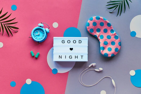 Puesta plana, caja de luz con texto "Buenas noches". Noche saludable sueño concepto creativo. Máscara para dormir, reloj despertador de menta azul, auriculares y tapones para los oídos. Fondo bicolor rosa y plateado con círculos de papel . —  Fotos de Stock