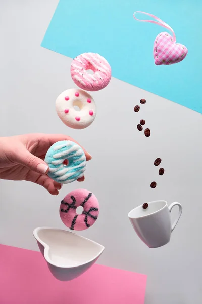 St. Valentine concept, levitation of doughnuts above heart shaped bowl. Coffee beans fly in espresso cup. Hand hold blue doughnut. Creative background in pastel shades of pink, blue and silver grey .