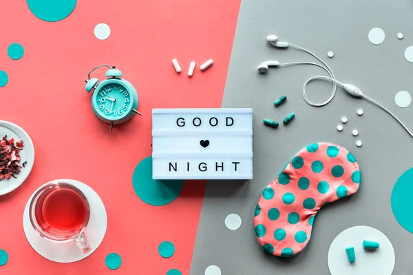 Texto "Buenas noches" en lightbox. Máscara de dormir rosa en lunares verdes azulejo, despertador, auriculares y tapones para los oídos. Colocación plana, vista superior, dividir dos tonos de coral naranja y fondo de papel plateado . —  Fotos de Stock