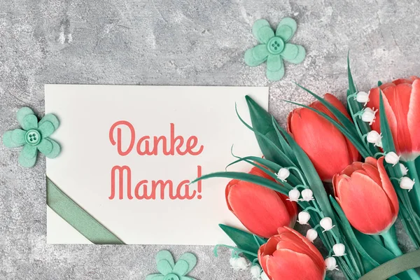 Данке Мама, німецьке привітання означає "Дякую, мама". Вітання в день матері на паперових картках з стрічкою. Різнокольорові тюльпани, текстильні квіти та паперові картки на сірому текстурованому тлі.. — стокове фото