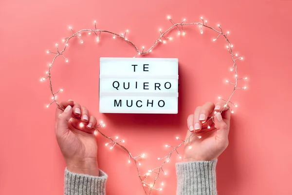 Valentine platte lay, bovenaanzicht op roze achtergrond. Lightbox met tekst "Te quiero mucho" betekent "Ik hou van je" in het Spaans. Lichte bloemenslinger in hartvorm in vrouwelijke handen. Valentijnsdag concept. — Stockfoto