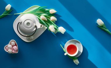Happy Valentine's or Mother's day celebration flat lay. Sweet marshmallow in heart shaped bowl.Tea cup, tea pot, sweets and white tulips on tale, background in trendy classic blue color.