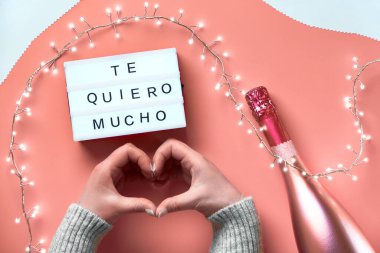 Valentine top view on pink background. Light garland, woman hands showing heart sign. Metallic pink champagne. Trendy monochrome flat layout . Text 