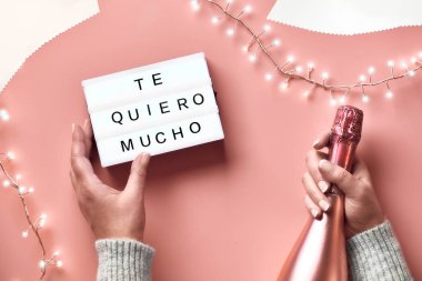 Valentine top view on pink background. Light garland, woman hands showing heart sign. Metallic pink champagne. Trendy monochrome flat layout . Text Te Quero Mucho means 
