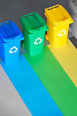 Three color coded recycle bins, isometric picture on grey paper, copy-space. Recycling sign on the bins, blue, yellow and green. Waste separation to minimize mixed waste and recycle paper and plastic.