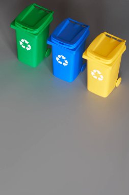 Three color coded recycle bins, isometric picture on grey paper, copy-space. Recycling sign on the bins, blue, yellow and green. Waste separation to reduce mixed waste and recycle paper and plastic.