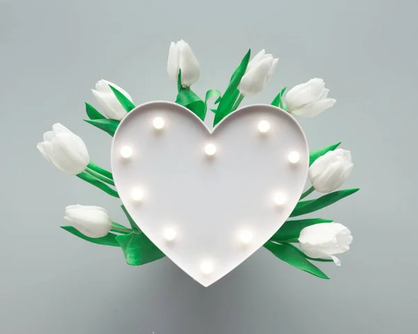 Spring background with lightboard in shape of heart illuminated with with lights. The heart is surrounded by white tulips on silver grey paper. Flat lay, top view from above, trendy springtime decor. — ストック写真