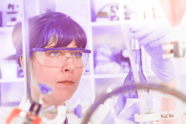 Young female scientist or tech in protective wear holds liquid biological sample, virus or flu vaccine. Coronavirus vaccine testing. Laboratory, neon purple light, blurred research facility.