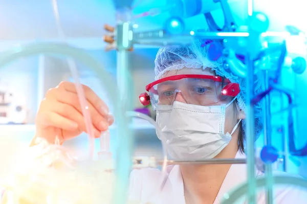Young female scientist or tech in protective wear supervises chemical synthesis, checking reaction. Laboratory, glassware, futuristic turquiose light, research facility out of focus.