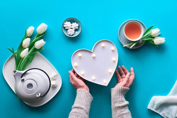 Spring geometric flat lay. Female hands show heart shape sign. Tea cup, tea pot, sweets and white tulips on blue table. Mothers day, international women day 8 March or your mom's birthday. — Stok fotoğraf