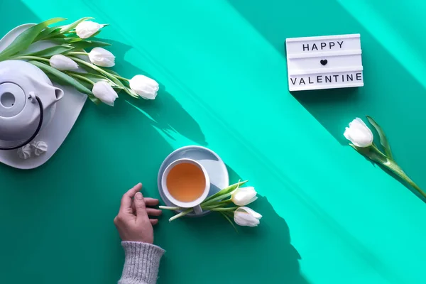 Wiosna geometryczna płaska leżała. Kobiece ręce trzymają skrzynkę z napisem "Szczęśliwych Walentynek". Filiżanka herbaty, dzbanek, słodycze i białe tulipany na zielono. Walentynki, światło słoneczne, długie cienie, nowoczesny design, widok z góry — Zdjęcie stockowe