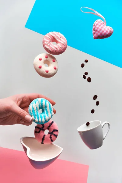 St. Valentine concept, levitation of doughnuts above heart shaped bowl. Coffee beans flying in espresso cup. Hand catching pink doughnut. Creative background in pink, blue mint and light grey color. — Stok fotoğraf