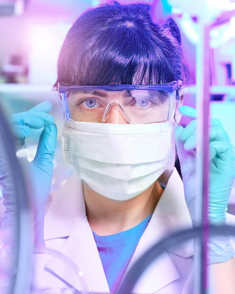 Young female scientist or tech in protective wear supervises chemical synthesis, checking reaction. Laboratory, glassware, futuristic neon light, research facility out of focus. — ストック写真