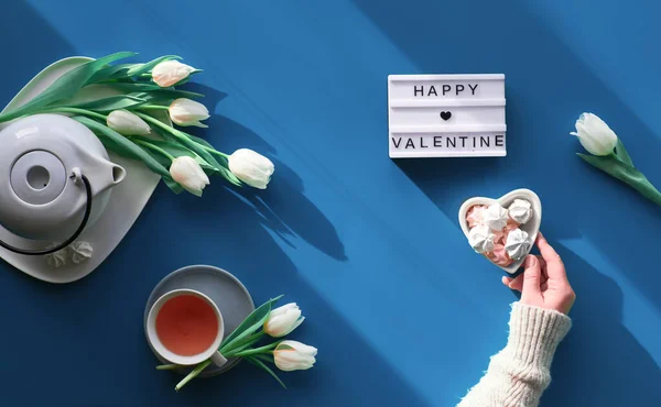 Happy Valentine's day celebration flat lay. Female hands show heart shape sign. Tea cup, tea pot, sweets and white tulips on tale, background in trendy classic blue color. — Stok fotoğraf